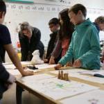 Students and elders stand around desks covered in artifacts, discuss objects.